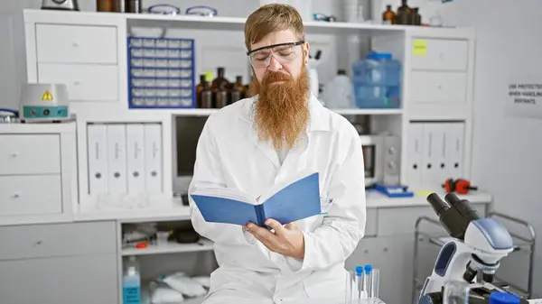Handsome young redhead man, serious scientist engrossed in reading research book, deep in thought at the heart of a buzzing laboratory.