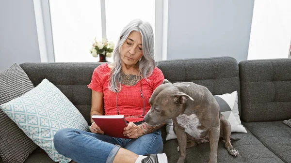 stock image Relaxed middle age woman, a grey-haired bibliophile, snuggled up with her loyal dog, comfortably sitting on her home sofa, indulging in the leisure of reading a riveting book in her cozy living room.