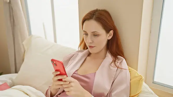 ピンクのローブで穏やかな白人女性がベッドでリラックスし 日光浴室でスマートフォンと婚約 — ストック写真