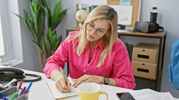 A focused blonde woman wearing glasses and a pink shirt is writing notes at her office desk, surrounded by office supplies.