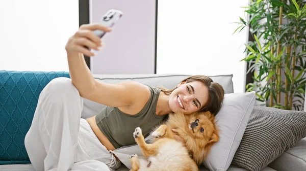 年轻而快乐的惊慌失措的女人舒舒服服地坐在自家客厅的沙发上 面带微笑 享受室内时光 用智能手机技术与宠物狗合影 — 图库照片