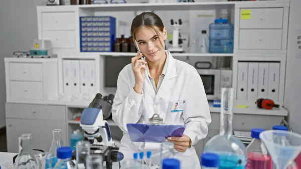 Stunning young hispanic woman scientist, reading report and talking on smartphone at lab work table, mastering the art of chemistry research