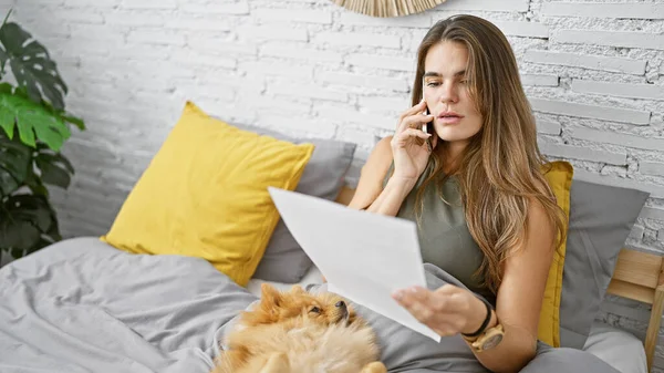 Relaxed morning talk, a confident young hispanic woman, smiling with her beloved dog in bed, is engaged in a lively conversation on her smartphone while comfortably reading a paper