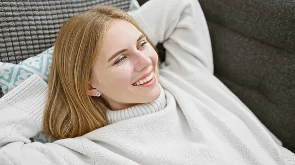 A smiling blonde woman with blue eyes wearing a white sweater relaxing comfortably in a cozy home interior.