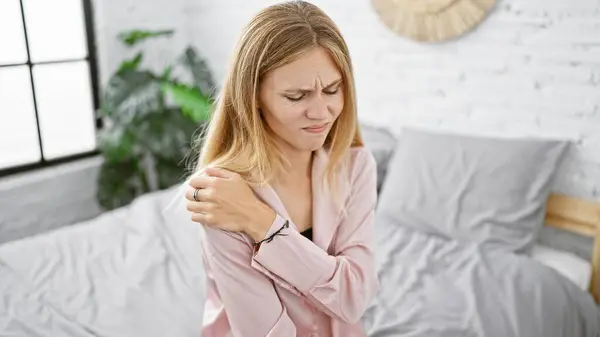 Blonde woman expressing pain while holding her shoulder in a bright bedroom setting