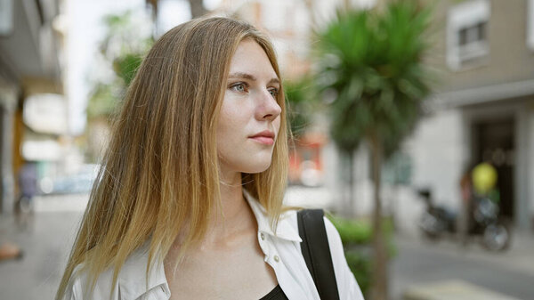 A thoughtful young woman with blonde hair and blue eyes gazes into the distance on a city street.