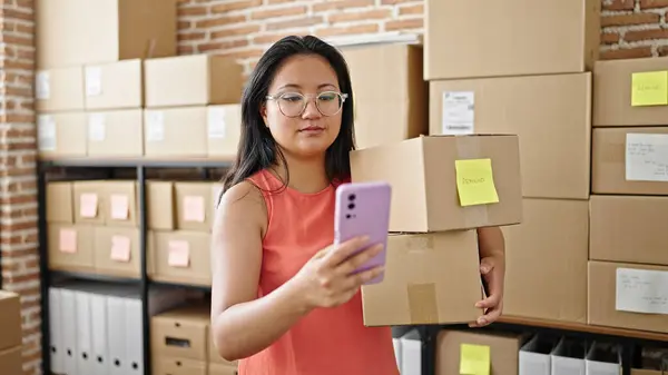 Young chinese woman ecommerce business worker using smartphone holding packages at office