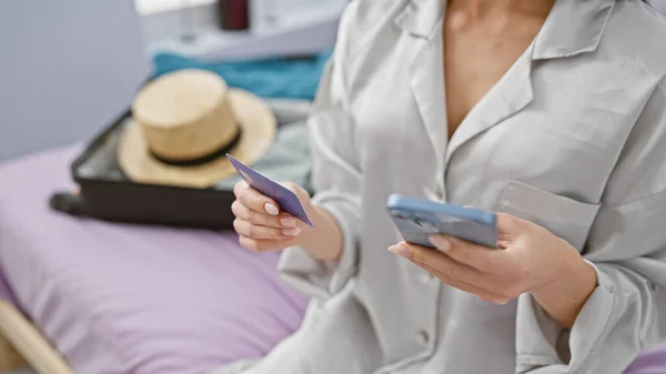 Beautiful young hispanic woman sitting in hotel room, using smartphone and holding credit card, surrounded by suitcase packed for vacation