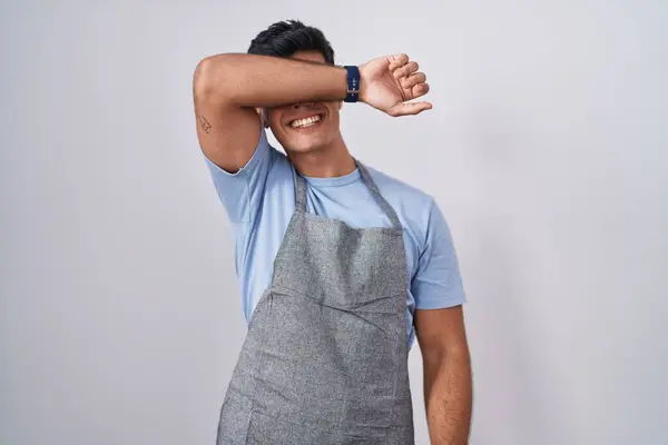 Hispanic young man wearing apron over white background covering eyes with arm smiling cheerful and funny. blind concept.