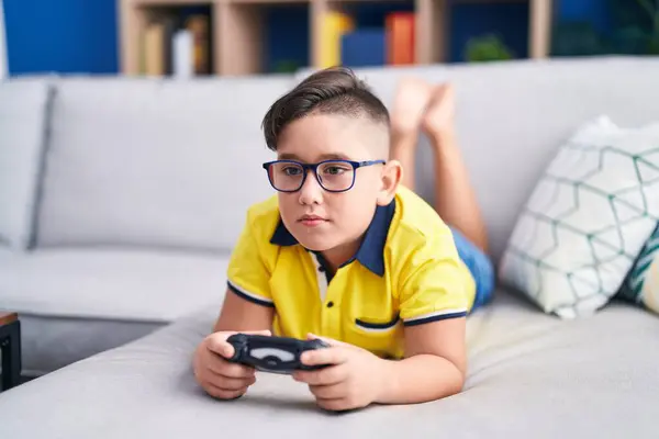 ソファーのコントローラーを保持するビデオゲームをプレイする若いヒスパニックの子供は 顔に深刻な表情でリラックスしました シンプルで自然なカメラを見る — ストック写真
