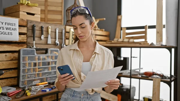 Young beautiful hispanic woman carpenter using smartphone reading document at carpentry