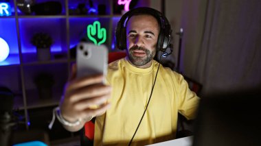 Sarı kazaklı, gülümseyen sakallı bir adam geceleri neon ışıklı bir oyun odasında selfie çeker..