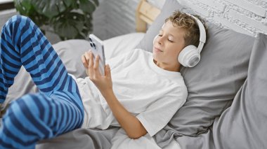 Sevimli sarışın çocuk, battaniyeye sarılmış, rahat bir odada bir yatakta dinleniyor, kendini kaptırmış ve akıllı telefonuyla video izlemeye odaklanmış. Sabahın erken saatlerinde, çocukların yatak odası..