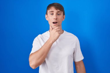 Mavi arka planda duran beyaz tenli sarışın adam elleri çenesinde, inanamama, şaşkınlık ve şaşkınlık dolu bir ifade takınıyor. 