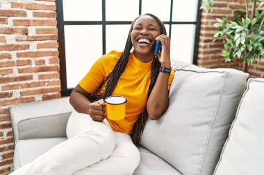 Afro-Amerikalı kadın akıllı telefondan konuşuyor evde kahve içiyor.