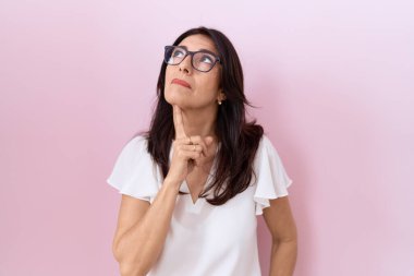 Orta yaşlı İspanyol bir kadın, günlük beyaz tişört ve gözlük takıyor. Düşünceleri kuşkuya odaklanmış, parmakları çenesinde ve yukarı bakıyor. 