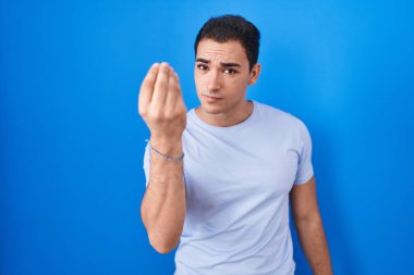 Young hispanic man standing over blue background doing italian gesture with hand and fingers confident expression 