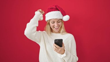Genç sarışın kadın noel şapkası takıyor. Akıllı telefon kullanıyor. Kırmızı arka plan üzerinde kazanan yüz ifadesi var.