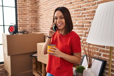 Young beautiful hispanic woman talking on smartphone drinking coffee at new home