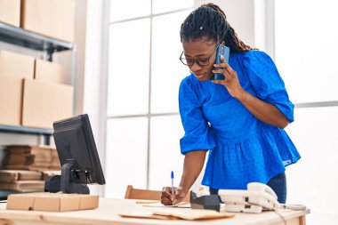 Afro-Amerikan kadın ekommerce iş işçisi ofisteki defterde akıllı telefonlardan bahsediyor.