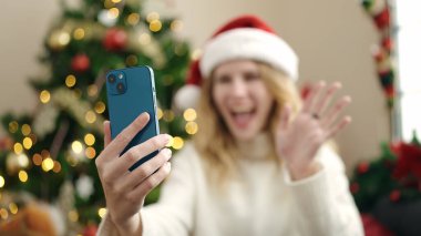 Young blonde woman having video call sitting on sofa by christmas tree at home