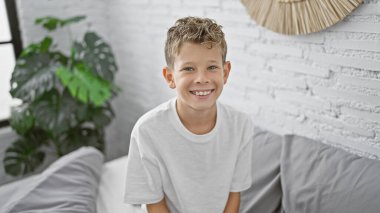 Sevimli sarışın çocuk, kendine güveni tam, güneşli bir yatak odasında rahat bir yatakta mutlu bir şekilde oturuyor, evde rahatlatıcı bir sabah geçiriyor.