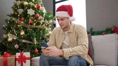 Genç İspanyol adam akıllı telefon kullanıyor. Yılbaşı ağacının yanında oturuyor.