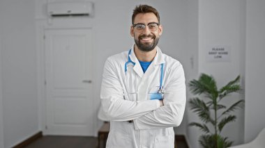 Kendine güvenen genç İspanyol bir adam, klinik bekleme odasında kolları çapraz duran gülümseyen bir doktor, pozitif cazibe ve profesyonel sağlık becerisi yayıyor.