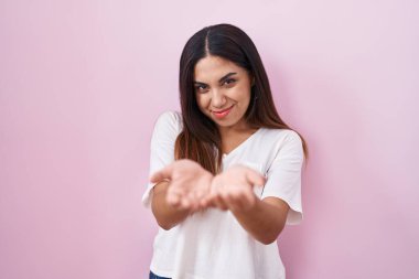 Pembe arka planda duran genç Arap kadın avuçlarını birbirine uzatarak gülümsüyor ya da el hareketi yapıyor. Bekleme ve koruma 