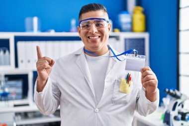 Laboratuvarda çalışan Latin asıllı genç adam elinde kimlik kartıyla gülümsüyor. 