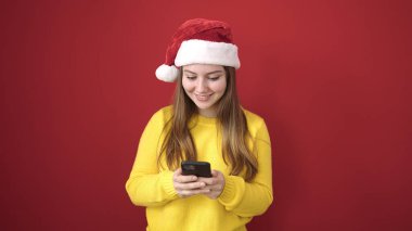 Yılbaşı şapkası takan genç sarışın kadın izole edilmiş kırmızı arka plan üzerinde akıllı telefon kullanıyor.