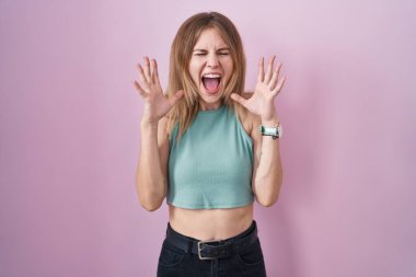 Blonde caucasian woman standing over pink background celebrating mad and crazy for success with arms raised and closed eyes screaming excited. winner concept 