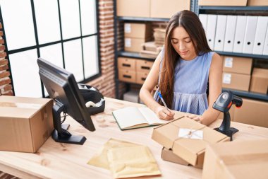 Young beautiful hispanic woman ecommerce business worker using computer writing on notebook at office