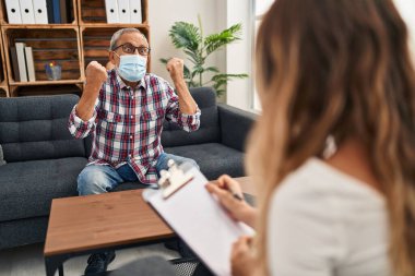 Neşeli kıdemli bir adam, tıbbi bir maske takıyor, bir psikoloji kliniğinde kollarını kaldırıp zaferinin ve heyecanının tadını çıkarıyor.!
