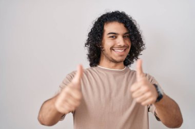 Beyaz arka planda duran kıvırcık saçlı İspanyol adam elle olumlu jest yapmayı onaylıyor, gülümsüyor ve başarı için mutlu. kazanan jest. 