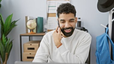 Modern ofis ortamında cep telefonuyla konuşan sakallı, gülümseyen genç bir İspanyol.