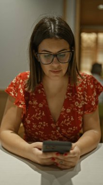 Kafe masasında akıllı telefon kullanan güzel İspanyol kadın, gözlüklü kendinden emin bir kadının portresi, içeride oturuyor, rahat ama odaklanmış.