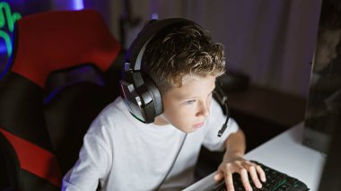 Sevimli sarışın erkek striptizci, fütüristik bir video oyunu oynamaya dalmış, gece dijital oyun odasından teknolojik aletler kullanarak canlı yayın yapıyor, yüzü odaklanmış ama rahat.