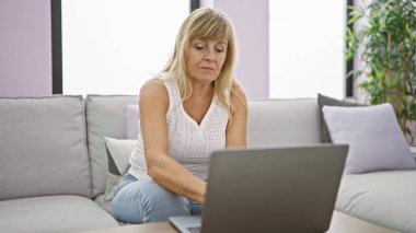 Konforlu evinde, ciddi ve odaklanmış orta yaşlı sarışın bir kadın koltukta oturuyor, tamamen içine kapanık, oturma odasındaki bilgisayarını kullanıyor..