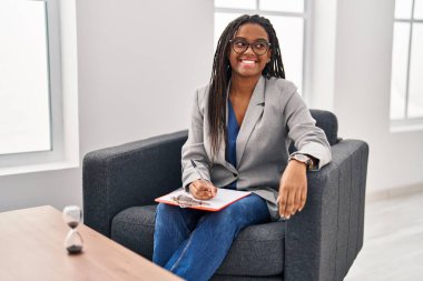 Danışmanlık ofisinde örgü ören genç Afro-Amerikalı yüzünde gülümseme ve doğal ifadeyle yan yana bakıyor. Kendinden emin gülen. 