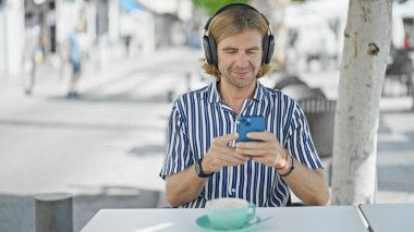 Sarışın, orta yaşlı bir adam, şehir merkezindeki bir kafede akıllı telefon kullanarak kulaklıkla müzik dinliyor..