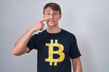 Bitcoin tişörtü giyen beyaz sarışın adam, el ve yüz parmaklarını ve burnunu göstererek neşeli bir şekilde gülümsüyor. güzellik konsepti 
