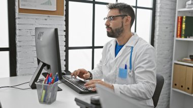 Sakallı, genç, İspanyol bir doktor, bir klinikte bilgisayarını kullanıyor.