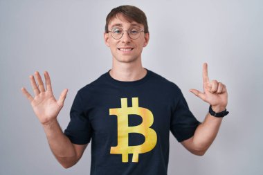 Bitcoin tişörtlü, beyaz, sarışın bir adam 7 numaralı parmaklarını gösterip gösterirken, kendinden emin ve mutlu bir şekilde gülümsüyor.. 