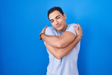 Mavi arka planda duran genç İspanyol adam kendine sarılıyor mutlu ve pozitif, kendinden emin gülümsüyor. Sevgi ve özgüven. 