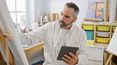 Parlak bir sanat stüdyosunda tablet kullanan gri saçlı, olgun sakallı bir adam..