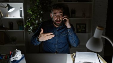 Genç, konsantre olmuş İspanyol adam gece yarısı lambasının parıltısıyla ofis masasında laptopuyla çalışırken akıllı telefonuyla ciddi bir konuşma yaptı.