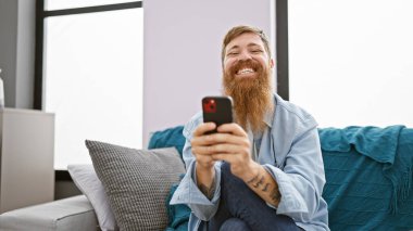 Yakışıklı genç kızıl saçlı adam evinin oturma odasındaki kanepede otururken neşeli bir şekilde telefonuyla mesajlaşarak ve internetteki dünyayla neşe ve pozitif titreşimler yayıyor.
