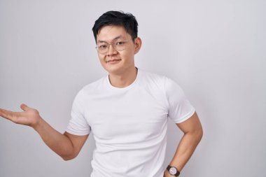 Beyaz arka planda duran Asyalı genç adam mutlu bir şekilde sunum yapıyor ve avuç içi kameraya bakıyor.. 