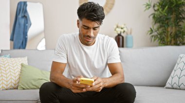 Yakışıklı İspanyol adam rahat bir oturma odasında akıllı telefon kullanıyor.